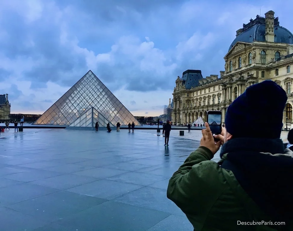 Turista tomando foto de la piramide del louvre, con un cielo de invierno bastante dramático
